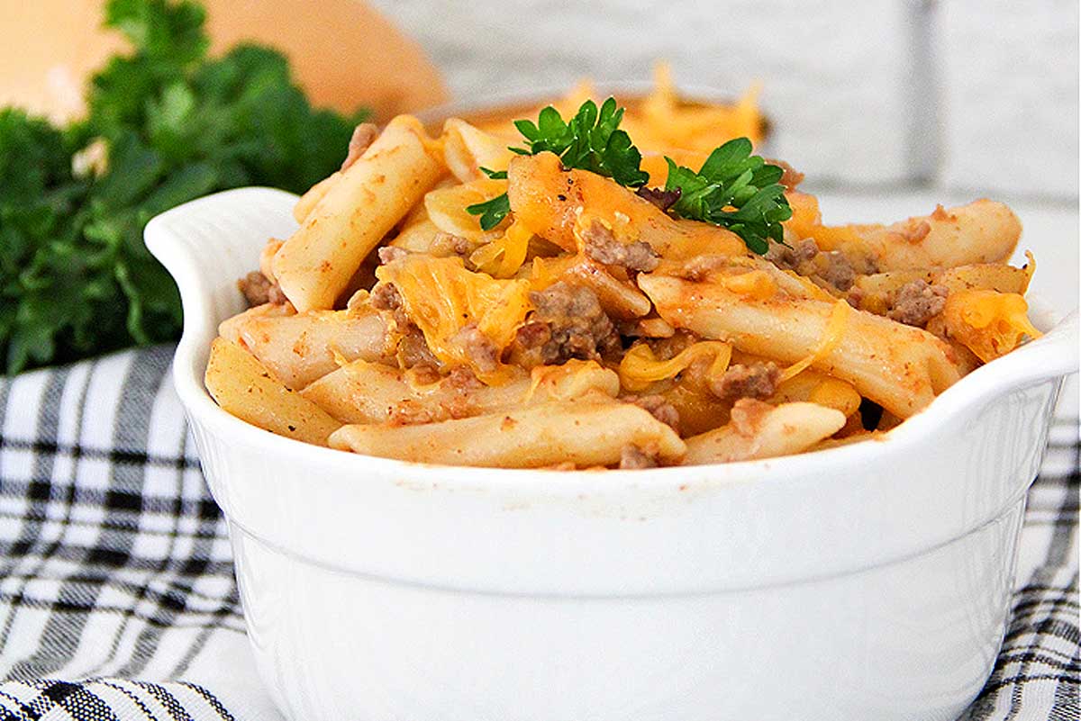 A white dish of baked ziti with melted cheese and meat, garnished with ketchup and parsley, on a checkered napkin.