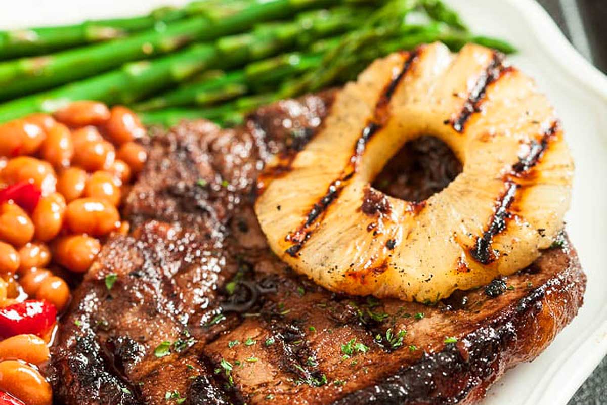 Grilled pork chop with a side of asparagus, baked beans, and a grilled pineapple ring on a white plate.
