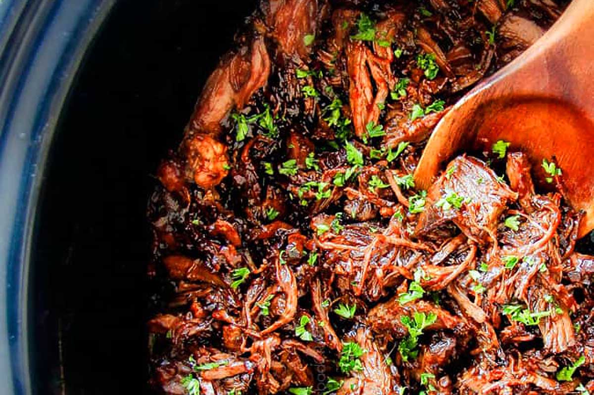 Slow-cooked pulled beef in a slow cooker, garnished with chopped parsley, with a wooden spoon.