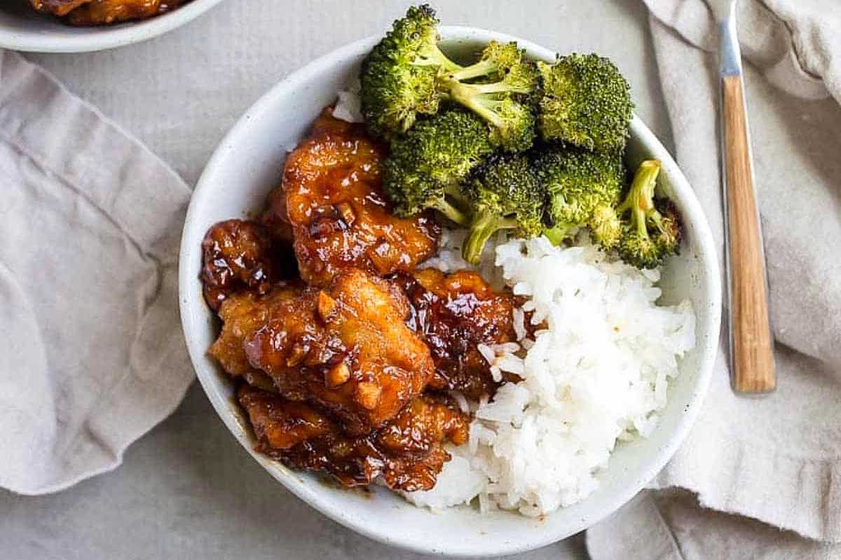 A bowl of orange chicken with broccoli and white rice, garnished with sesame seeds and ketchup, presented on a light gray surface.