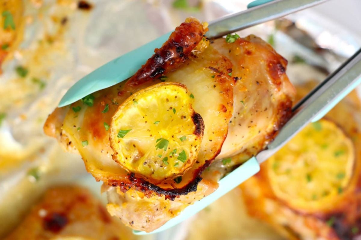 Close-up of metal tongs holding a piece of roasted chicken topped with a slice of lemon and garnished with chopped herbs, perfect for those exploring delicious lemon recipes.