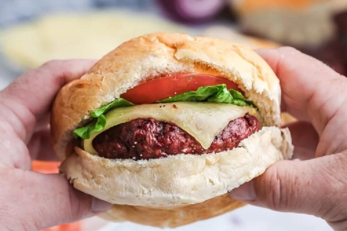 Hands holding a burger crafted from ground beef recipes, with cheese, lettuce, and tomato slice in a bun.