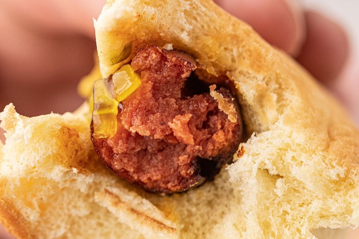Close-up of a bitten sandwich with a spicy, smoked sausage patty and pickles inside a white bun.