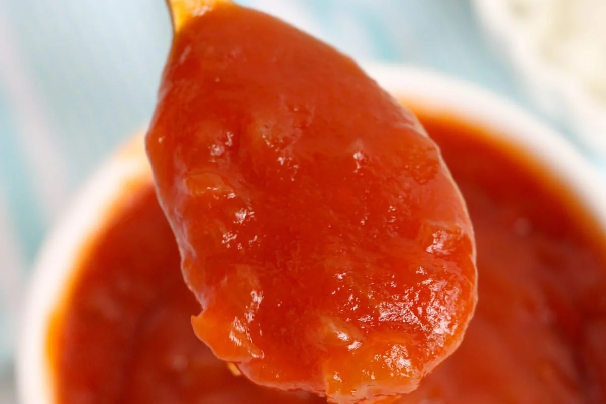 A spoonful of thick, red tomato ketchup held above a bowl filled with more ketchup, perfect for trying out your favorite ketchup recipes.