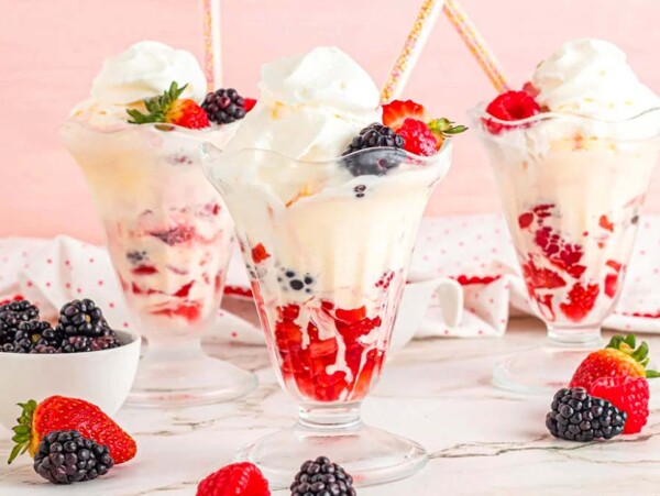 Three fruity desserts: ice cream sundaes topped with whipped cream and mixed berries in elegant glass cups, with extra berries scattered around.