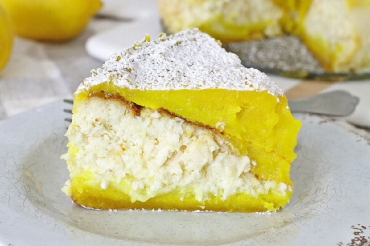 A slice of lemon ricotta cake dusted with powdered sugar graces a white plate, epitomizing the allure of classic lemon recipes, with a fork artfully positioned in the background.
