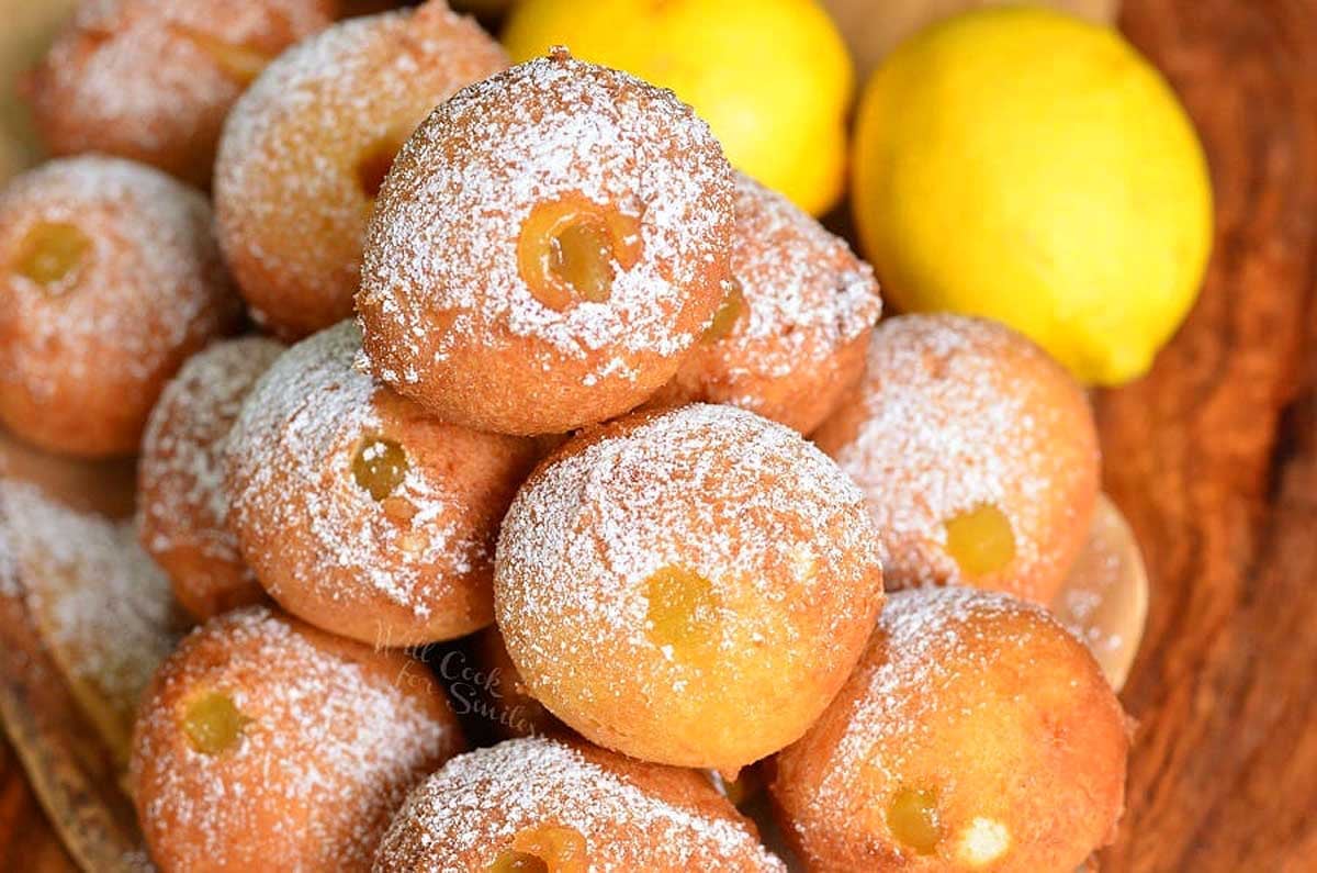 A pile of lemon curd-filled doughnuts dusted with powdered sugar, arranged on a wooden serving tray with fresh lemons in the background.