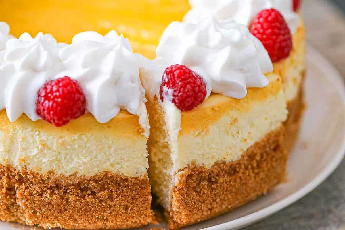 A slice of creamy cheesecake with a graham cracker crust, topped with lemon curd and raspberries, served on a white plate.