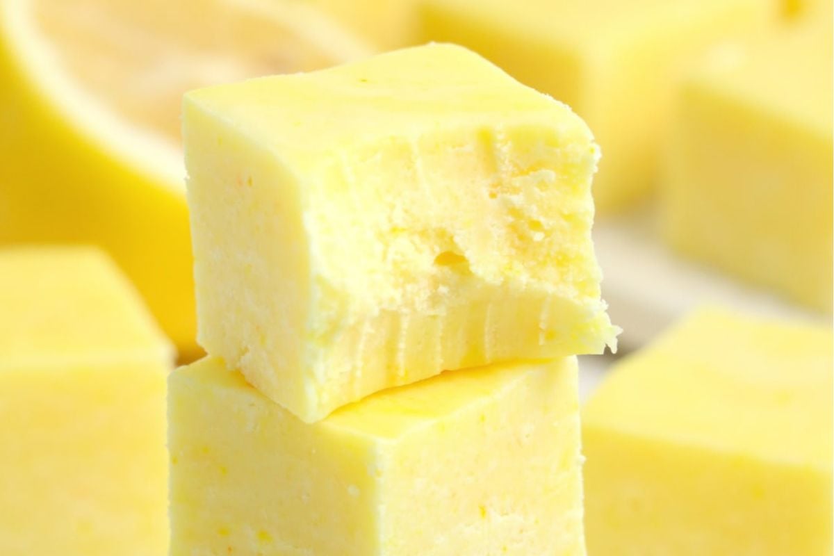 Two creamy yellow lemon fudge squares, one with a bite taken out, stacked with more in the background—an irresistible treat for anyone who loves lemon recipes.