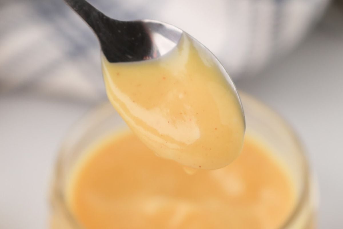 A spoonful of creamy yellow sauce, possibly mustard or dressing, held above a jar of the same sauce. A blurred background with a striped cloth is visible, perfect for brightening up your favorite lemon recipes.