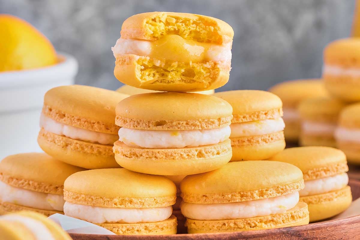 A stack of yellow macarons with a bite taken out of the top one, revealing a lemon curd filling, placed on a wooden surface.