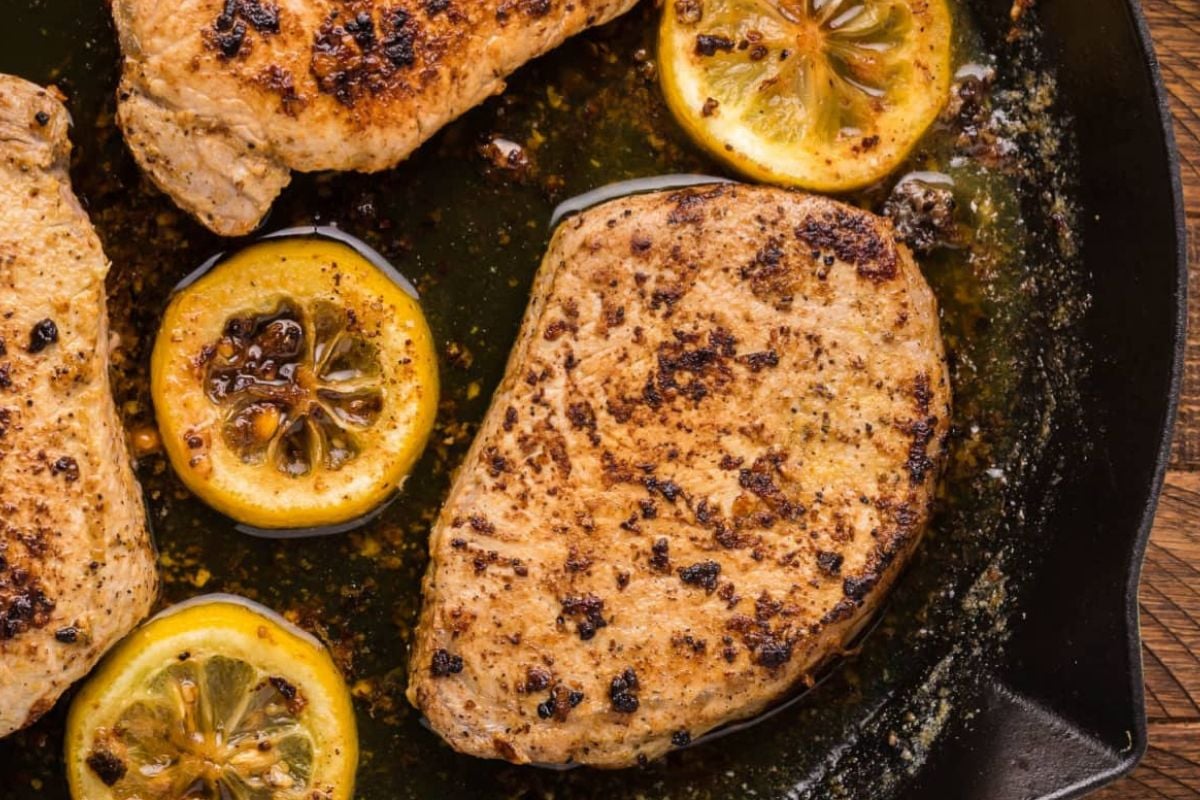 Seared pork chops with lemon slices in a skillet, topped with spices and herbs—a perfect addition to your favorite lemon recipes.