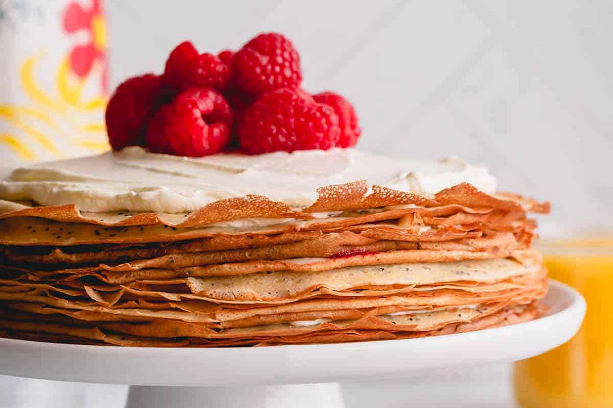A stack of thin, golden crepes layered with lemon curd, topped with fresh raspberries on a white cake stand against a softly blurred background.