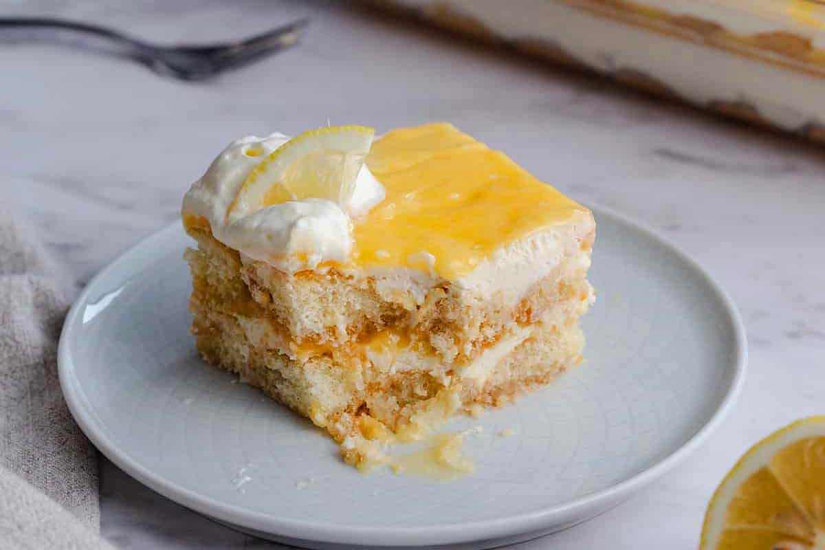 A slice of lemon cake topped with whipped cream and a lemon curd garnish, served on a white plate with a fork in the background.