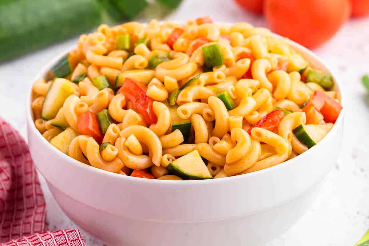 A bowl of colorful pasta salad with chopped vegetables, including green cucumbers and red tomatoes, on a kitchen counter with a red napkin and a side of ketchup.