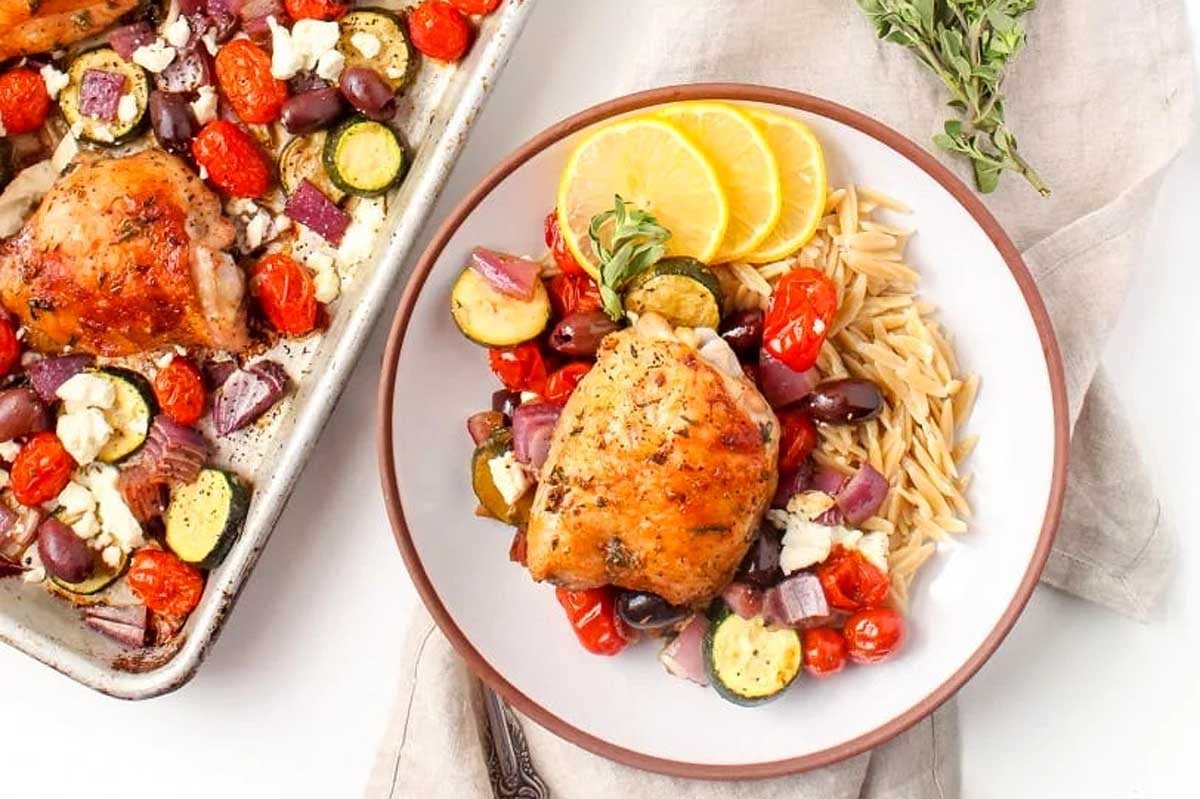 A plate of roasted chicken thigh served with orzo pasta and a side of sheet pan roasted Mediterranean vegetables, garnished with lemon slices and fresh herbs.