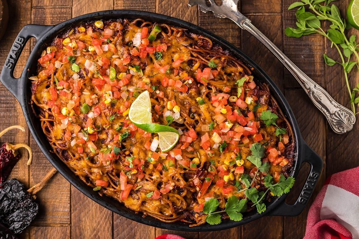 A sizzling cast-iron skillet filled with enchiladas topped with melted cheese, fresh tomato salsa, and lime wedges, accompanied by recipes featuring noodles.
