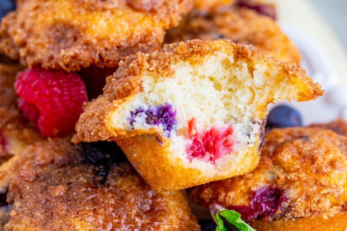 A close-up of freshly baked mixed blueberry muffins with a focus on a half-eaten muffin revealing the colorful berries inside.