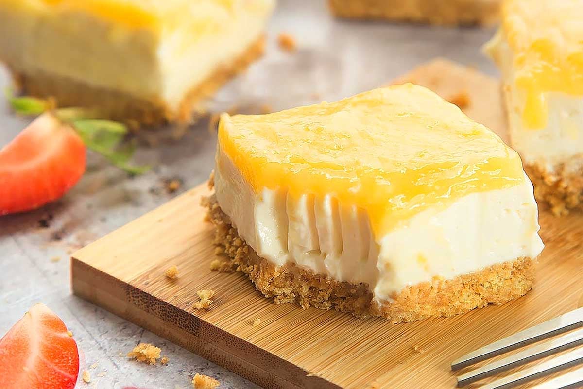 A slice of creamy cheesecake with a lemon curd topping and crumbly base, served on a wooden board with a fork and strawberry slices nearby.