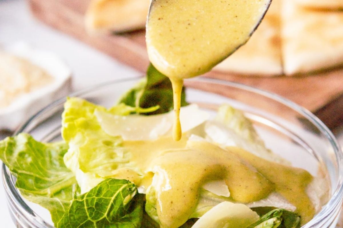 A spoon is drizzling creamy dressing, inspired by lemon recipes, onto a fresh green salad in a clear glass bowl.