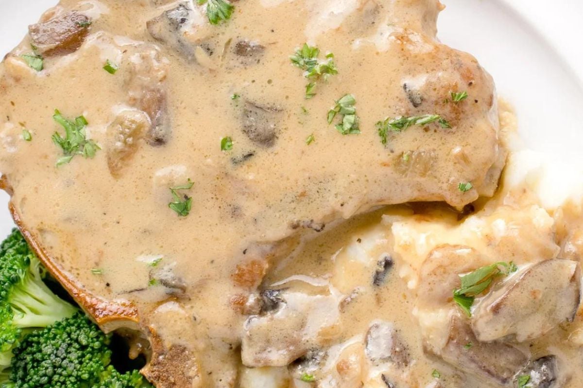A close-up of a pork chop recipe covered in mushroom sauce, served with mashed potatoes and broccoli on a white plate.