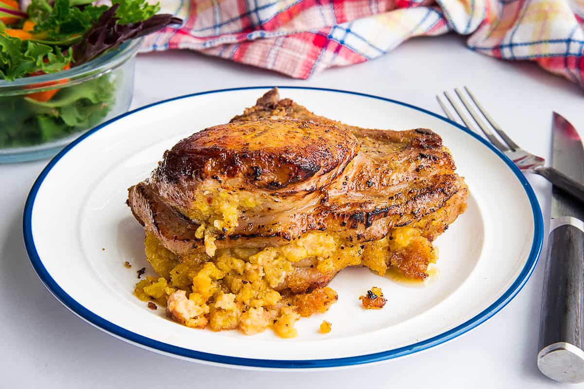 A roasted pork chop recipe on a white plate with stuffing, accompanied by a salad and a fork on a kitchen table.