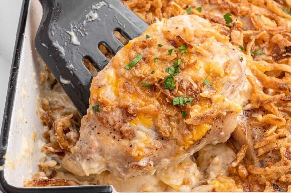 Close-up of a creamy chicken casserole made with a can of cream soup, topped with crispy onions, garnished with chopped chives, being served with a spatula.
