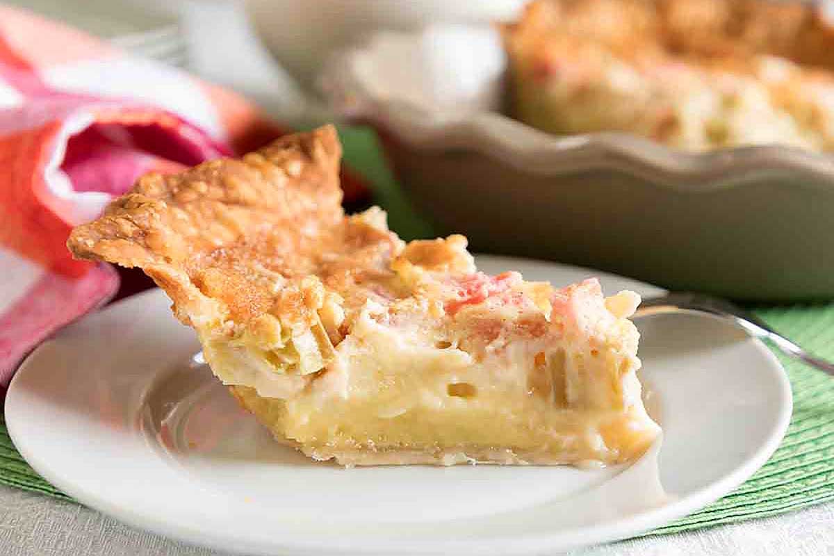 A slice of creamy seafood pie with visible chunks of crab and shrimp, served on a white plate, accompanied by a colorful rhubarb-infused napkin on the side.