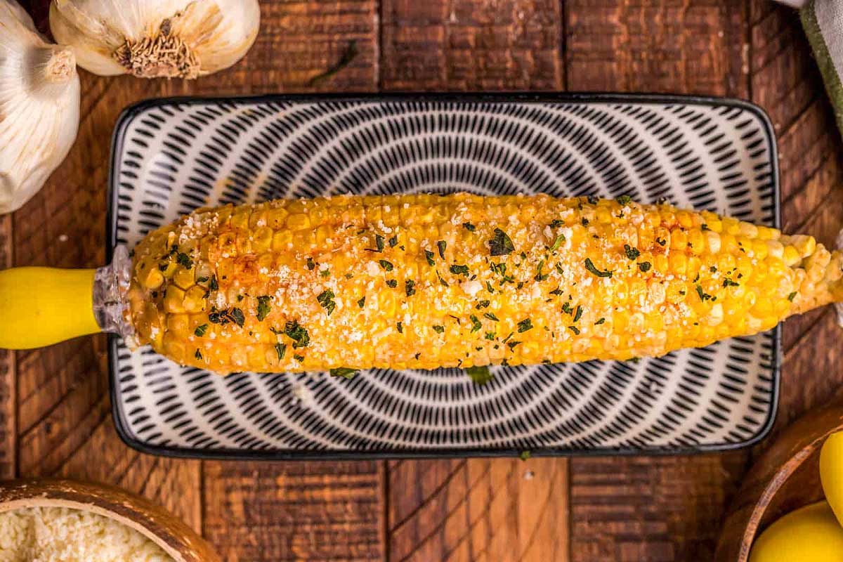 Smoked corn on the cob with butter, herbs, and spices, served on a decorative plate with garlic cloves in the background.