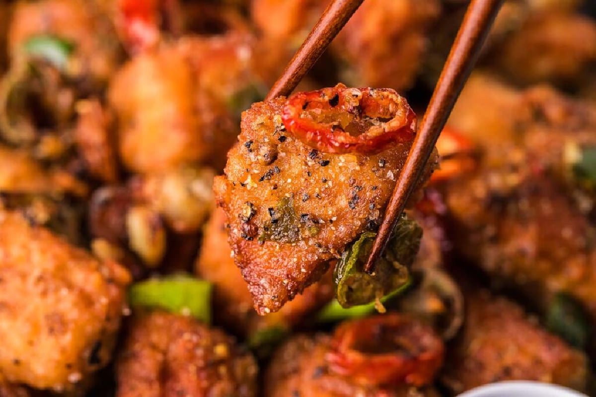 Chopsticks holding a piece of spicy fried pork chop with visible seasoning and chopped green onions, with more pork chops in the background.