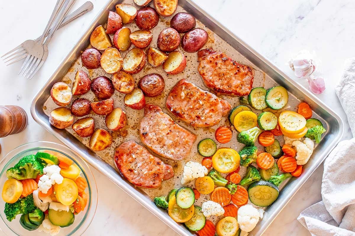 A sheet pan with roasted pork chop recipes, potatoes, and mixed vegetables including zucchini, carrots, and cauliflower, prepared and ready to serve.