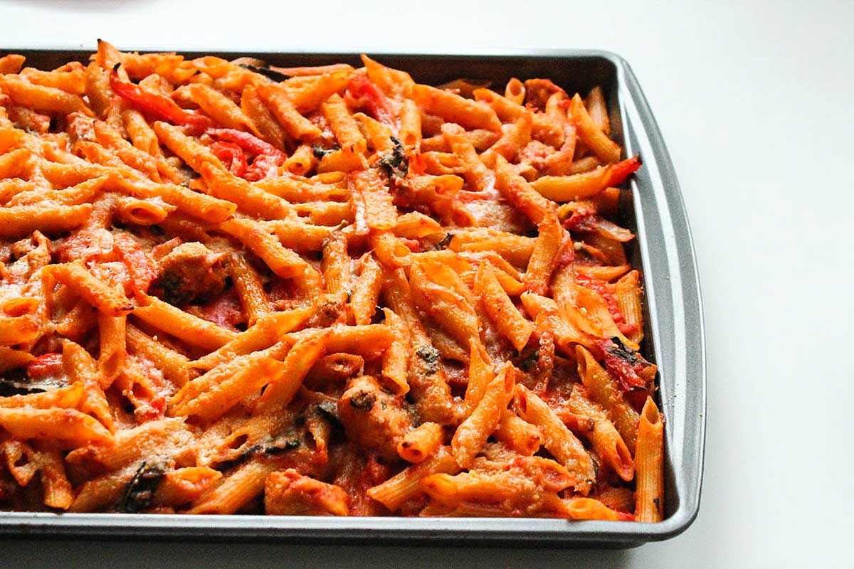 A sheet pan of baked penne pasta with tomato sauce and roasted vegetables on a white background.