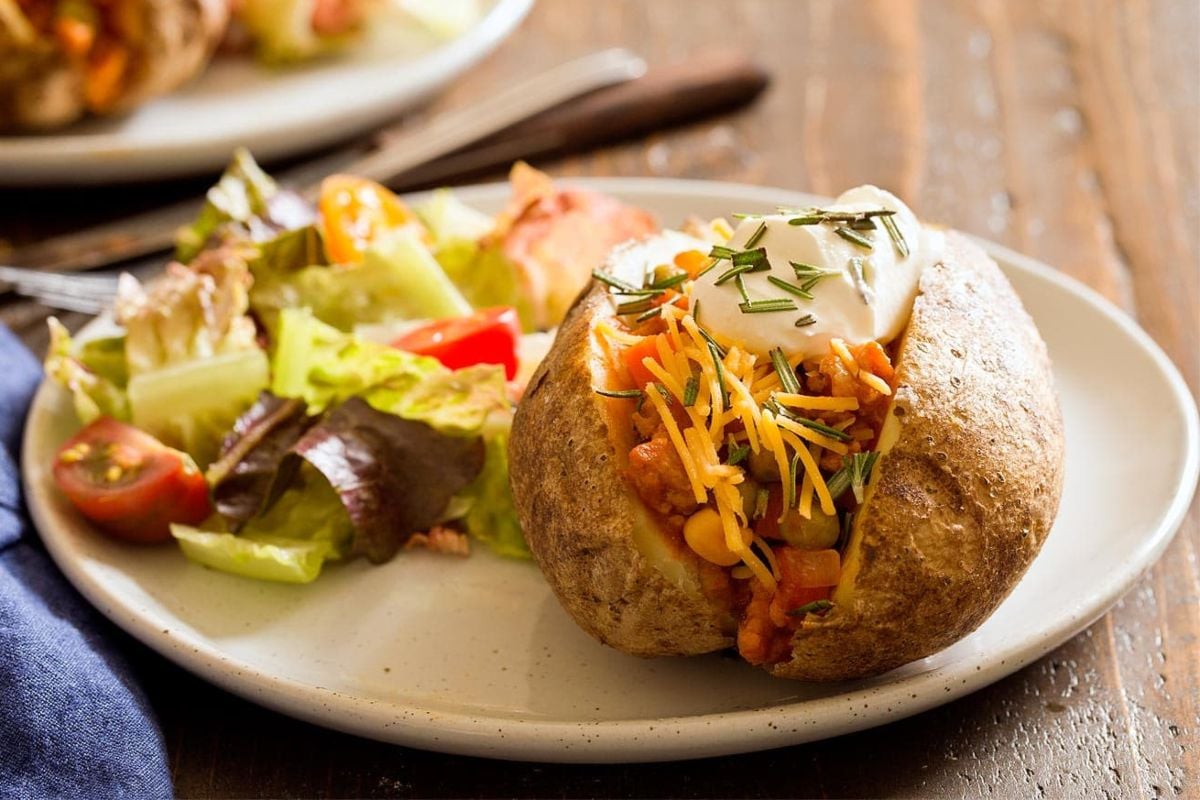 A baked potato topped with cheese, using frozen vegetables, sour cream, and herbs, served with a side salad on a white plate.