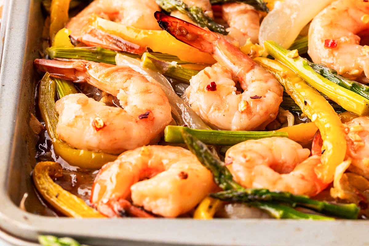 Cooked shrimp and sliced yellow bell peppers with asparagus in a sheet pan.