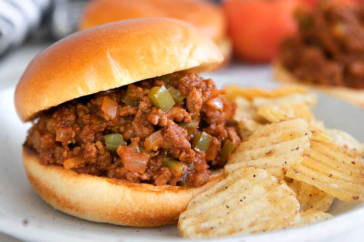 A ketchup-based sloppy joe sandwich on a white plate with crinkle-cut potato chips on the side.