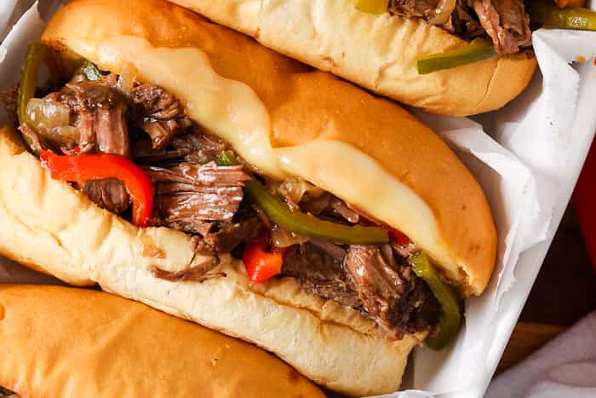 Three slow cooker philly cheesesteak sandwiches with sliced beef, melted cheese, and peppers in white takeout containers.