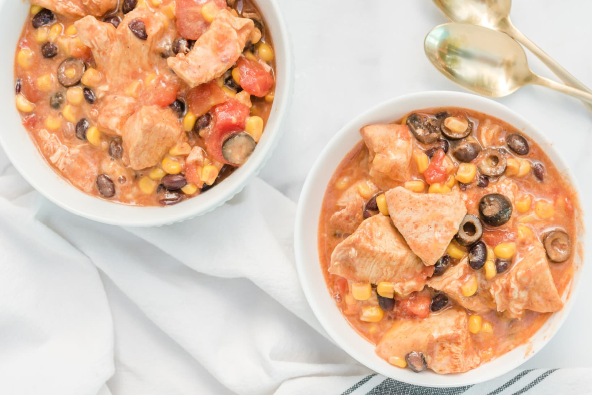 Two bowls of creamy chicken chili with corn, black beans, olives, and tomatoes, beside two gold spoons—a comforting choice for Slow Cooker Dinners and Sides.