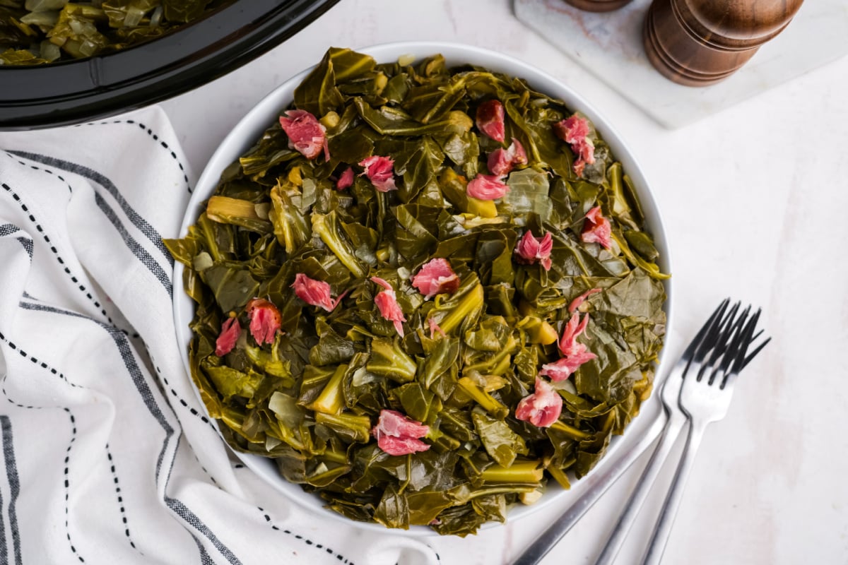 A bowl of cooked collard greens with pieces of ham, perfect for Slow Cooker Dinners and Sides, sits next to a striped towel and metal forks.