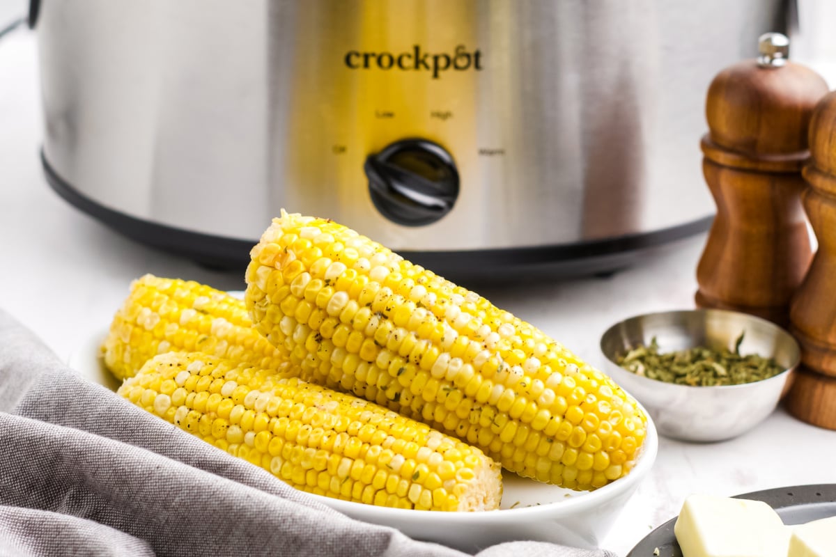 Three cooked corn cobs on a plate, perfect as Slow Cooker Dinners and Sides, with a Crockpot, herbs, salt, and pepper shakers in the background.