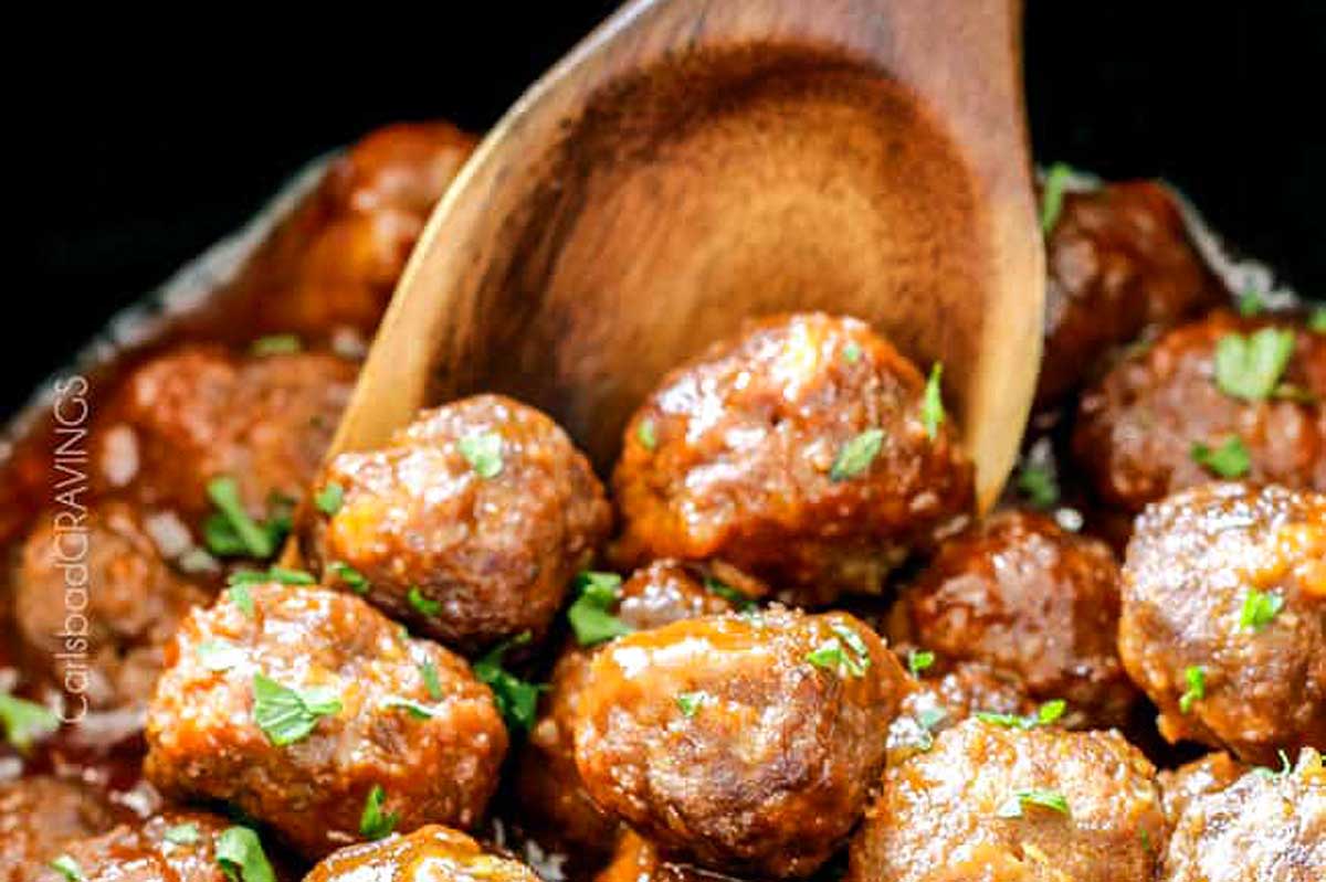A wooden spoon scooping glazed meatballs garnished with parsley from a slow cooker.