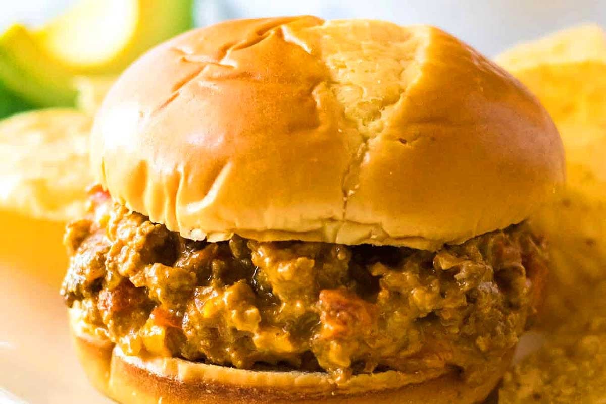 A close-up image of a sloppy joe sandwich on a bun, featuring Velveeta cheese, served with chips and a slice of lemon in the background.