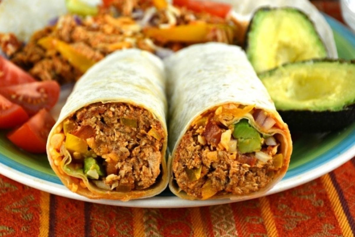 Two chicken burritos with a meat and vegetable filling served on a plate with sliced avocado and tomatoes on the side.