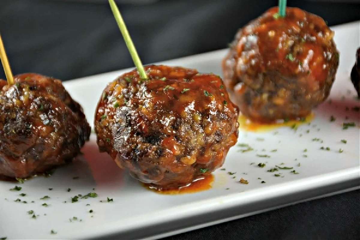 Meatballs covered in a tangy ketchup-based sauce on a white plate, each with a toothpick, garnished with chopped herbs—a delicious twist on classic ketchup recipes.