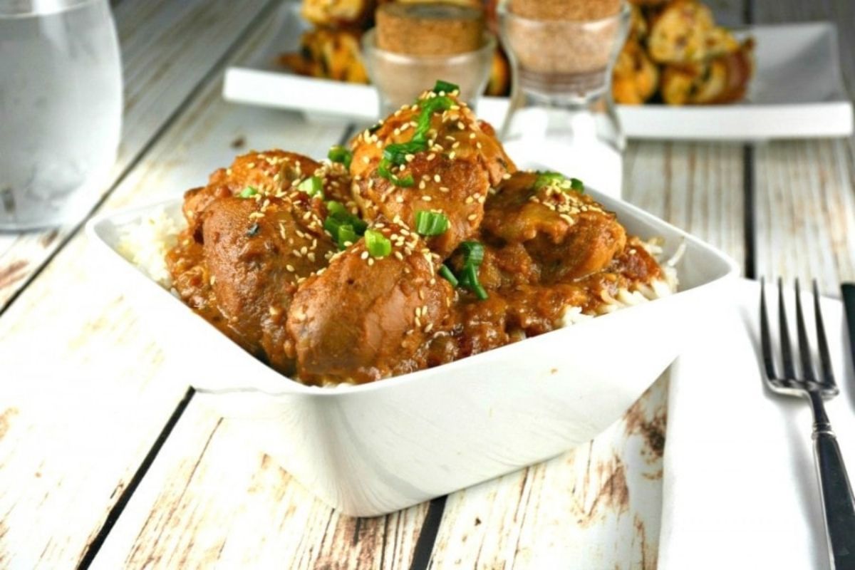 A bowl of sesame chicken dinners served over rice, with a fork on the side and additional food items in the background.