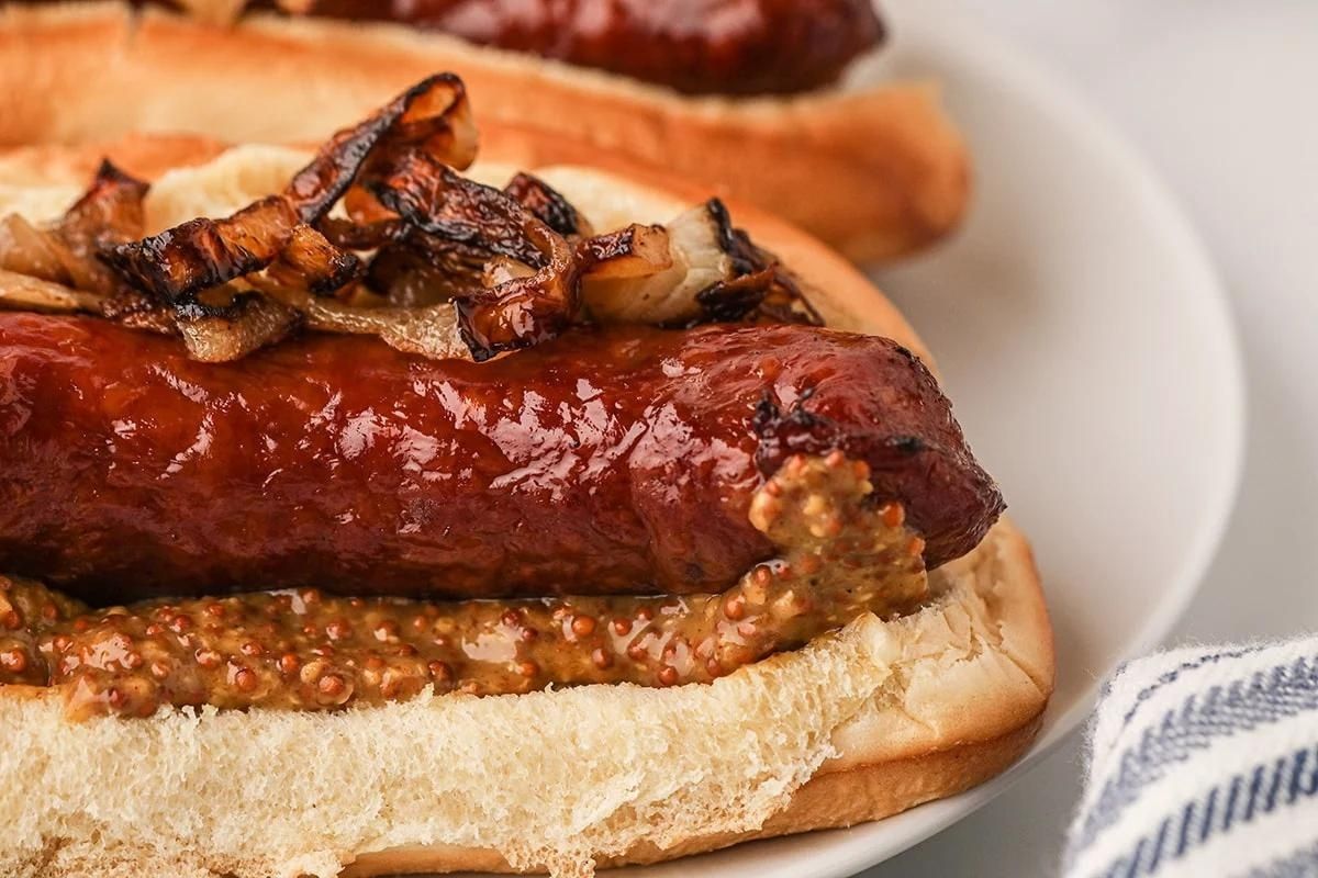 A hot dog with a shiny, grilled sausage topped with mustard and caramelized onions, served in a white bun on a plate.