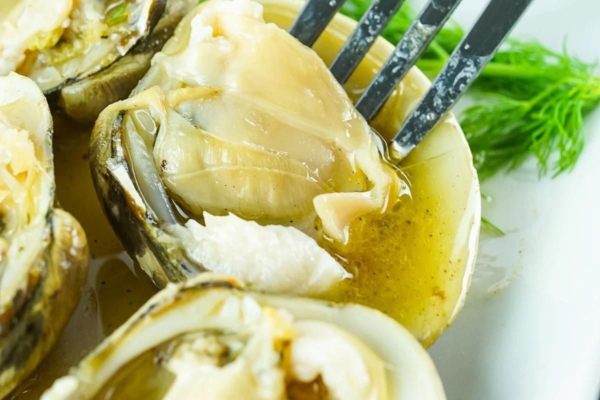 Close-up image of smoked clams on a plate with a fork lifting one, garnished with fresh dill, highlighting texture and juiciness.