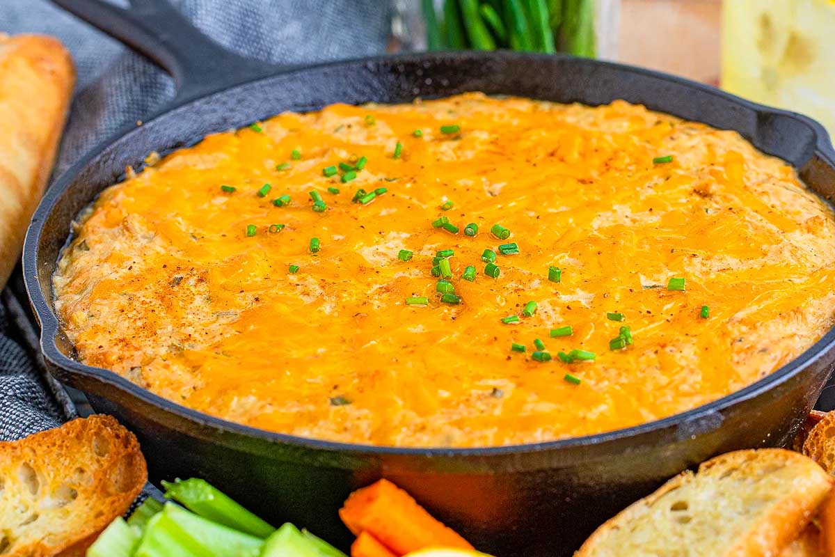 A skillet of creamy orange buffalo chicken dip topped with melted cheese and garnished with green onions, served with smoked bread and fresh vegetables on the side.