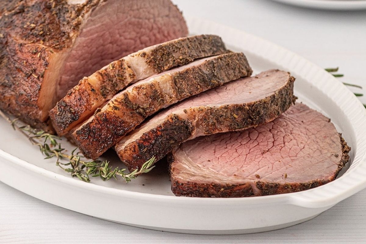 Sliced smoked roast beef on a white plate garnished with thyme, showcasing a pink center and a seasoned crust.