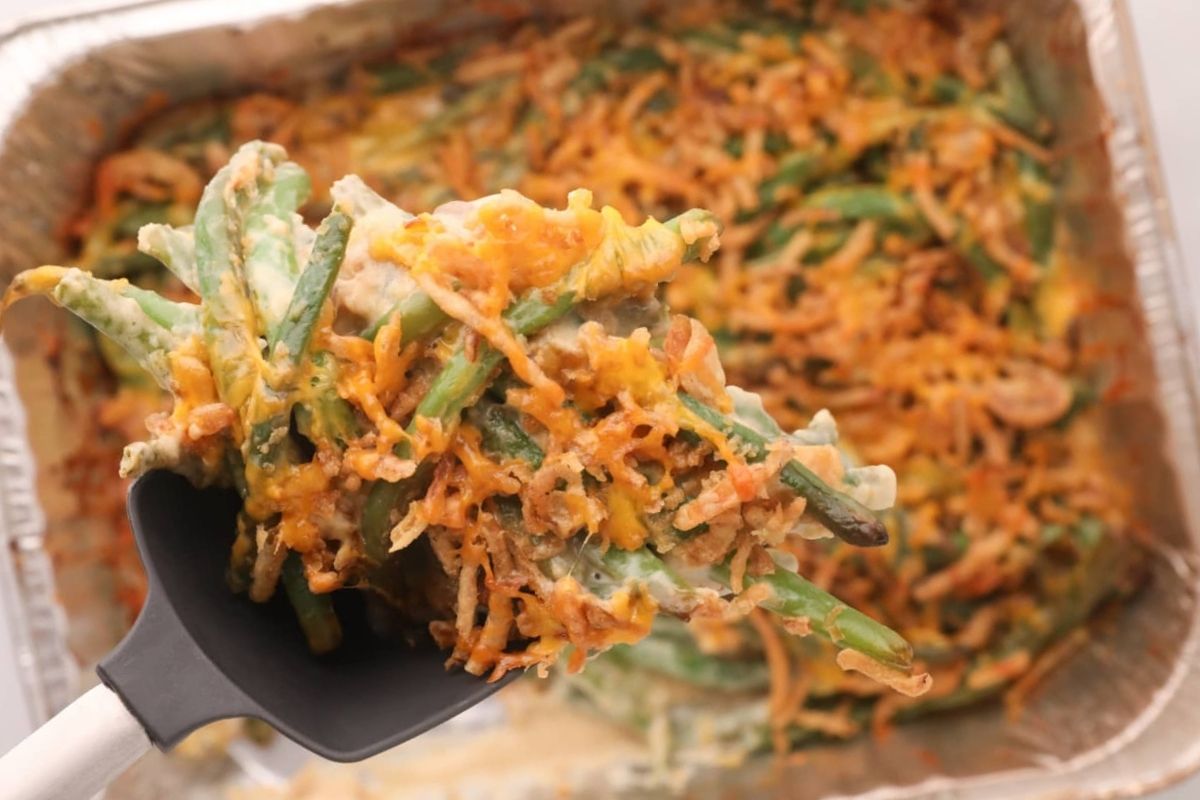 A serving spoon lifting green bean casserole with crispy smoked onions from a foil tray.