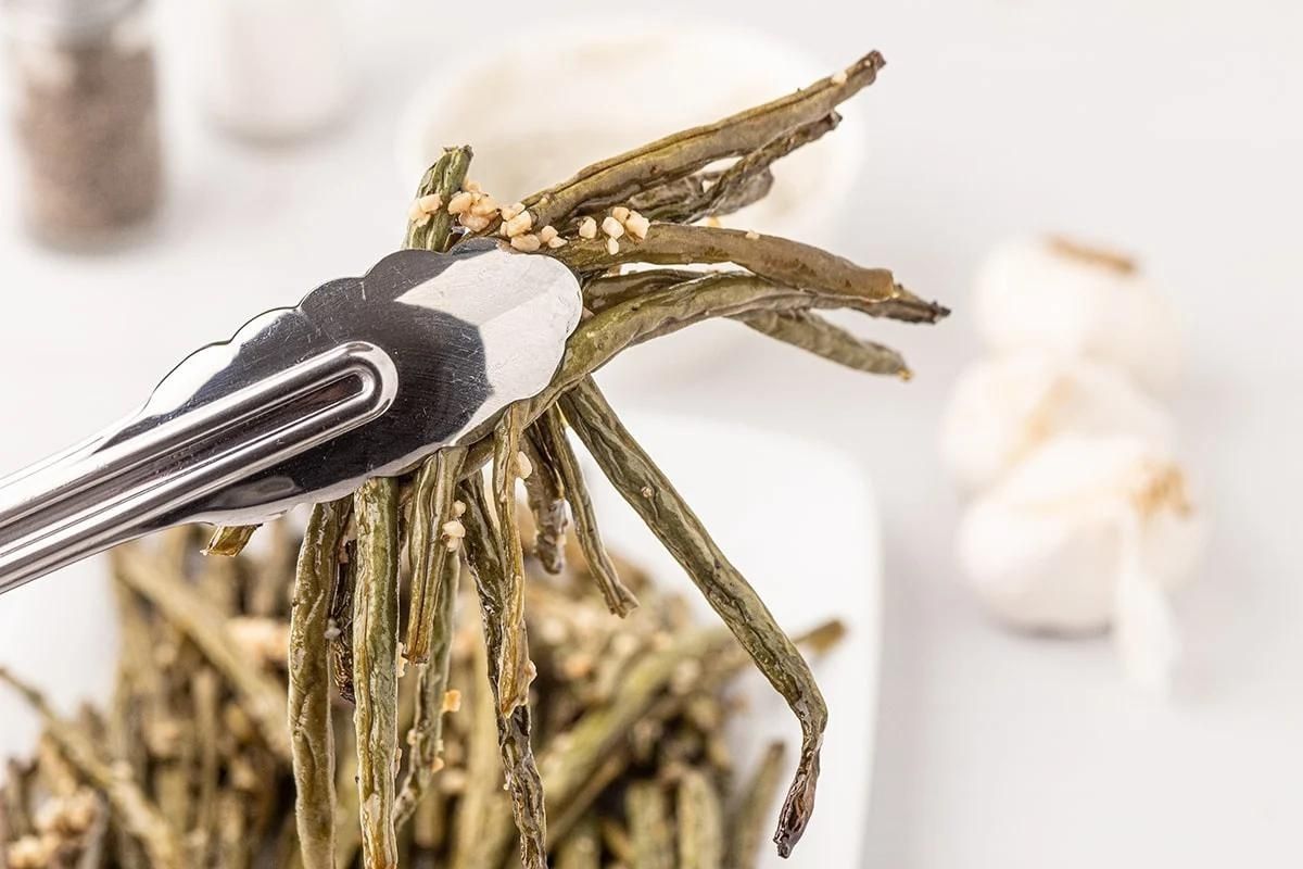 Tongs holding smoked asparagus spears with seasonings, with garlic cloves and condiments blurred in the background.